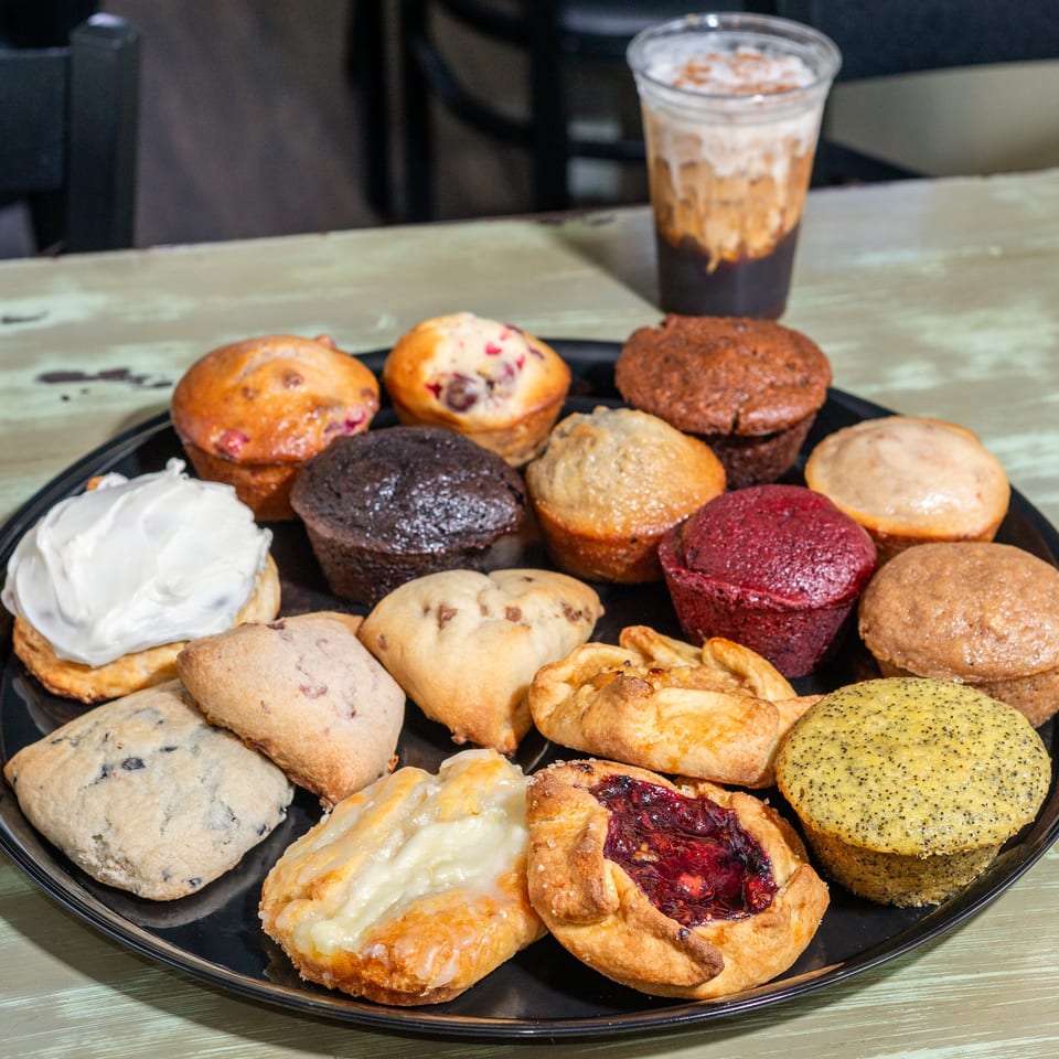 A black tray of assorted muffins, scones, and pastries with an iced coffee in the background.