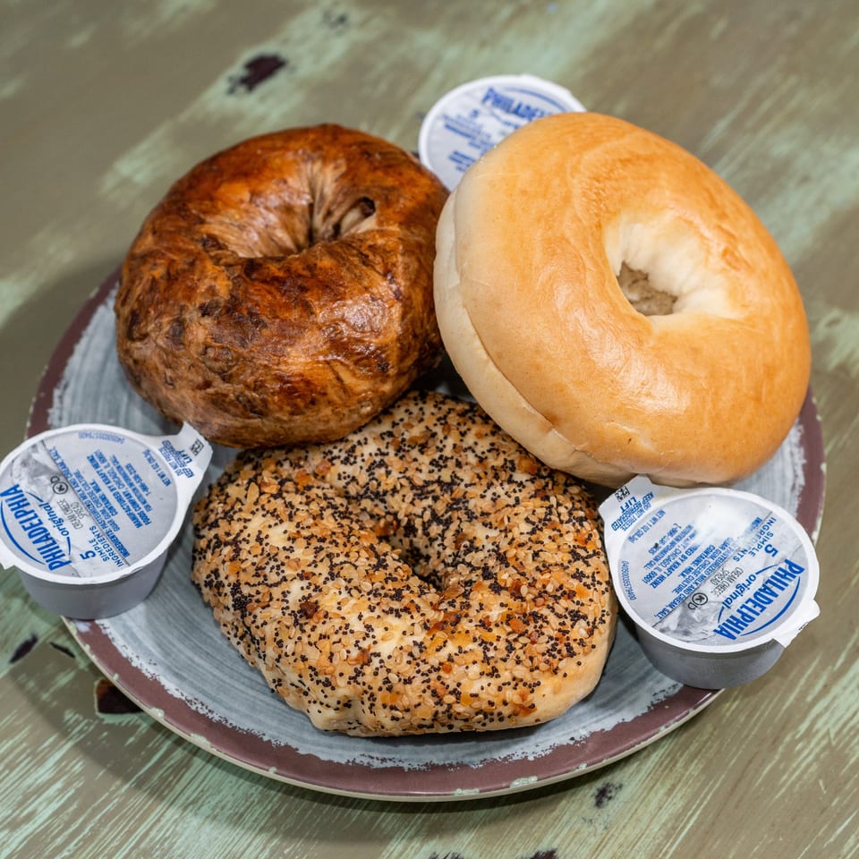 Three bagels, plain, cinnamon raisin, and everything, with four Philadelphia cream cheese cups on a plate.