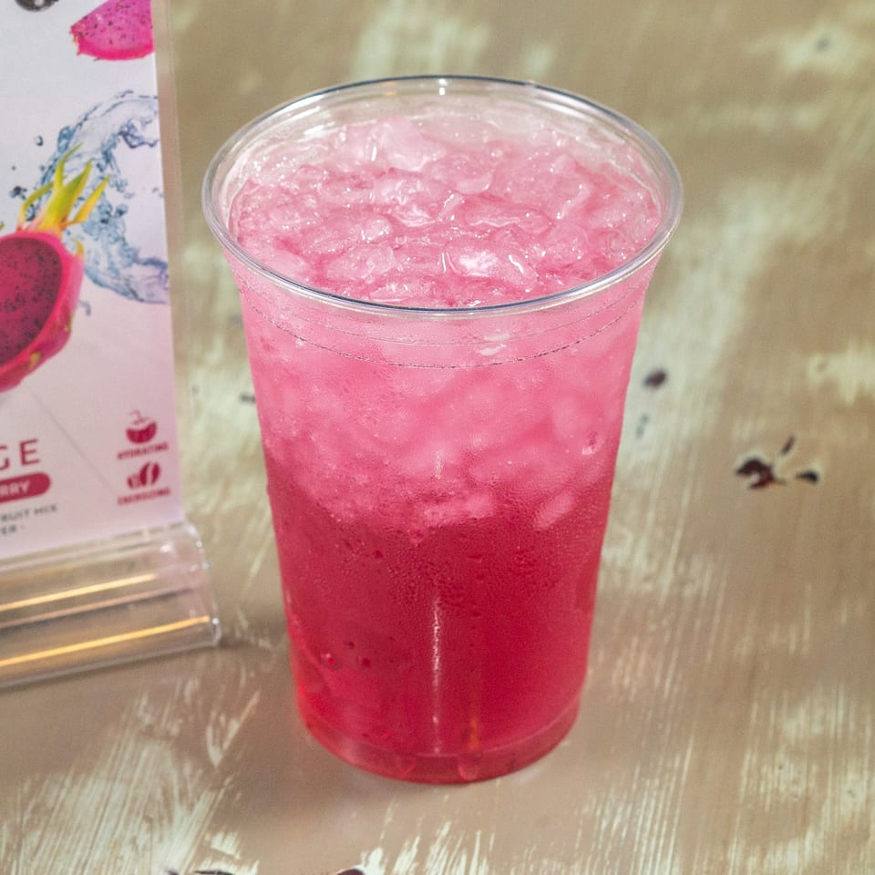 A vibrant pink iced drink in a clear plastic cup, with a menu featuring dragon fruit in the background.