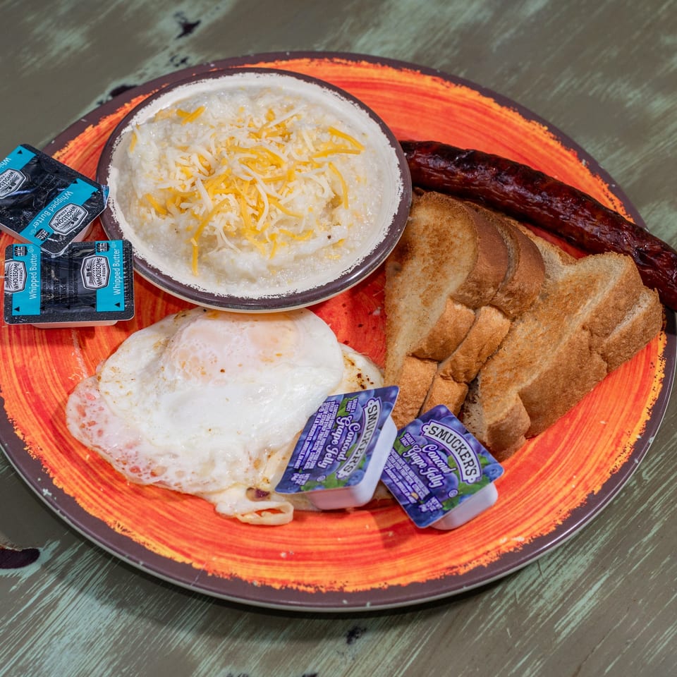 A breakfast plate with cheesy grits, fried eggs, sausage, toast, butter, and grape jelly.